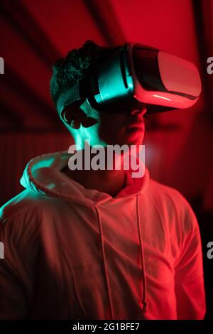 Uomo che indossa una felpa con cappuccio mentre usa occhiali VR e in piedi in studio con luci al neon rosse Foto Stock