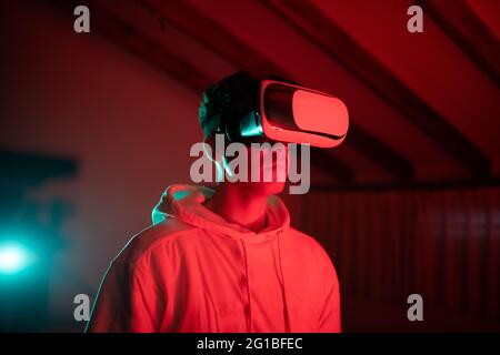Uomo che indossa una felpa con cappuccio mentre usa occhiali VR e in piedi in studio con luci al neon rosse Foto Stock