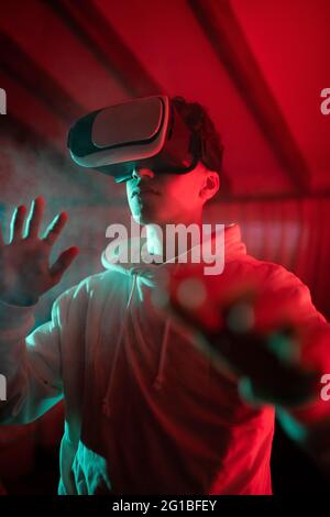 Uomo che indossa una felpa con cappuccio mentre usa occhiali VR e in piedi in studio con luci al neon rosse Foto Stock