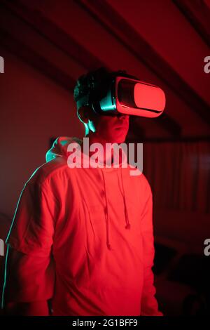 Uomo che indossa una felpa con cappuccio mentre usa occhiali VR e in piedi in studio con luci al neon rosse Foto Stock
