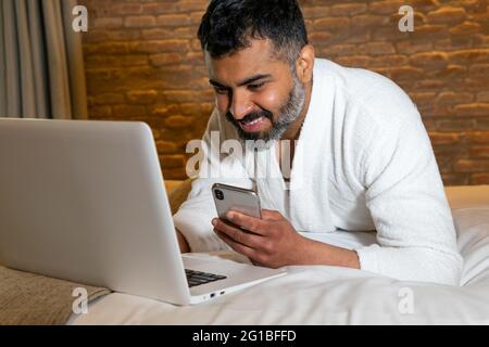 Crop sorridente adulto maschio in bianco accappatoio navigare netbook e telefono cellulare mentre si sdraia su morbido letto in camera d'albergo Foto Stock
