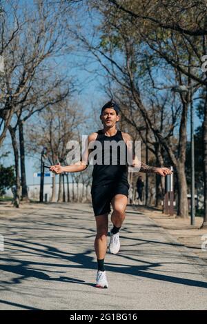 Atleta maschile in abbigliamento sportivo saltando con la corda saltante e guardando via sulla passerella durante l'allenamento cardio nel parco Foto Stock