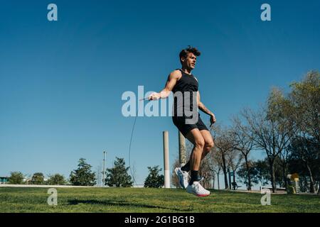 Atleta maschile in abbigliamento sportivo saltando con la corda saltante e guardando via sulla passerella durante l'allenamento cardio nel parco Foto Stock