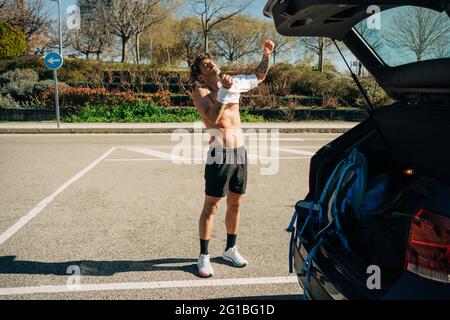 Atleta maschile maschile con tatuaggio che mette la t-shirt contro l'automobile in parcheggio il giorno di sole Foto Stock