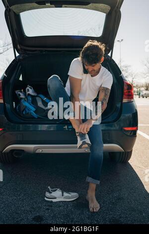 Atleta maschile tatuato che mette su scarpe mentre parla sul cellulare contro l'auto con tronco aperto in città Foto Stock