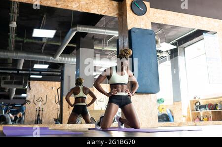 Atleta afroamericana in activewear con le mani sulle anche che respirano durante l'allenamento contro lo specchio in palestra Foto Stock