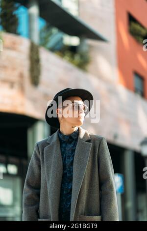 Giovane uomo transgender in cappotto e cappello di classe che guarda via alla luce del giorno Foto Stock