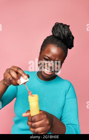 Donna afro-americana felice con gli occhi chiusi indossando abiti blu e soffiando bolle di sapone contro sfondo rosa in studio Foto Stock