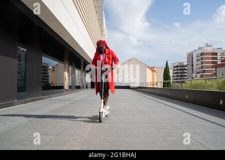 Corpo pieno di donna afroamericana in maschera con brecce brillanti che guidano scooter elettrico lungo la costruzione della torre a tempo libero Foto Stock