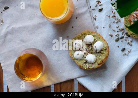 Dall'alto di toast di yummy con avocado e palline di mozzarella decorate con lino e semi di girasole e servite su tovagliolo vicino a bicchieri di arancio fresco Foto Stock