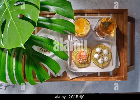Dall'alto di deliziosi toast di avocado con salmone e burrata servito a tavola con bicchieri di succo fresco e tè alle erbe posto vicino a Mo esotico Foto Stock