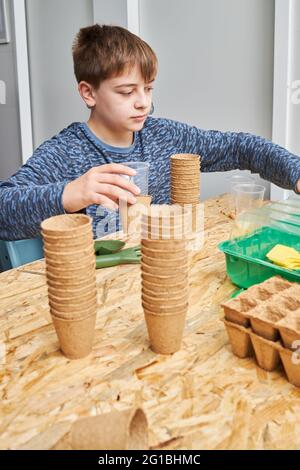 Bambino seduto al tavolo con pila di tazze di cartone e contenitori contro pala da giardinaggio e forchetta mentre si guarda via Foto Stock