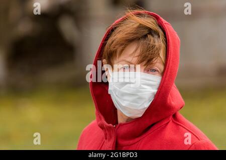 Una donna anziana con capelli marroni e occhi blu in un cappotto rosso e cappuccio in una maschera medica sicura protettiva, primo piano ritratto. Foto Stock