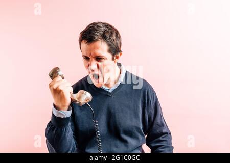 giovane adulto trentacinque anni uomo in camicia blu con collo a v e maglione con sfondo rosa arrabbiato urlando a un telefono con cavo Foto Stock