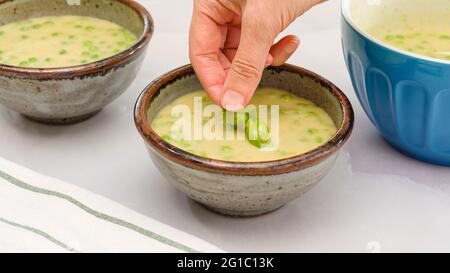 Zuppa cremosa di sedano al purè servita con foglie di basilico fresco da vicino in una ciotola sul tavolo da cucina, le mani della donna, lo sfondo in marmo chiaro Foto Stock