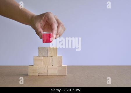 Mano mettendo un blocco di legno rosso su pila di blocchi piramidali di legno. Unicità o distinguersi dal concetto di pacchetto. Foto Stock