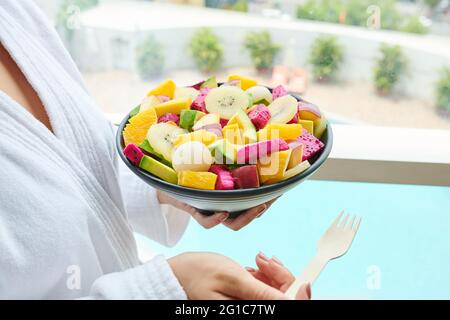 Giovane donna in accappatoio che mangia una deliziosa Macedonia di frutta per colazione e che guarda attraverso la finestra Foto Stock