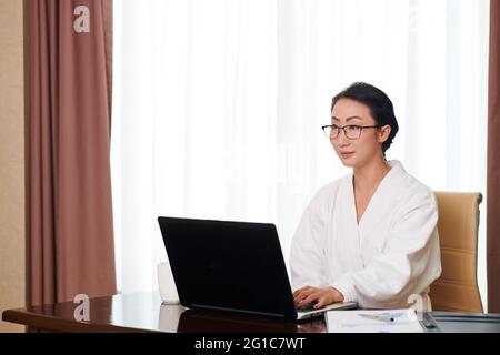 Ritratto di giovane bella donna d'affari in accappatoio e occhiali che lavorano su computer portatile a casa Foto Stock