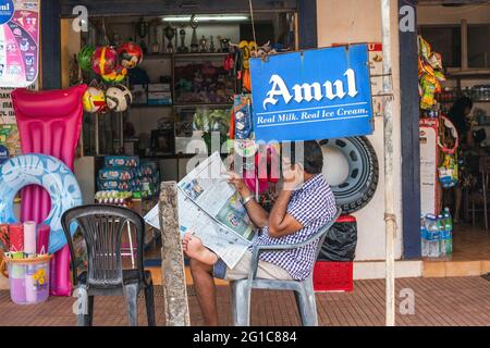 Indian Gentleman siede il giornale di lettura fuori del minimarket che vende gli accessori della spiaggia, Agonda, Goa, India Foto Stock