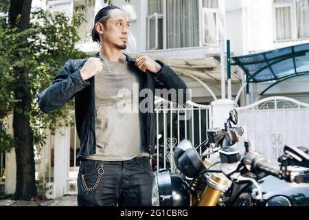 Motociclista serio e penoso in t-shirt sudata in piedi in bicicletta e indossare giacca nera in pelle Foto Stock