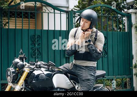 Uomo pensiver seduto su una motocicletta, indossando un equipaggiamento da guida e una maglietta, allacciando il casco Foto Stock