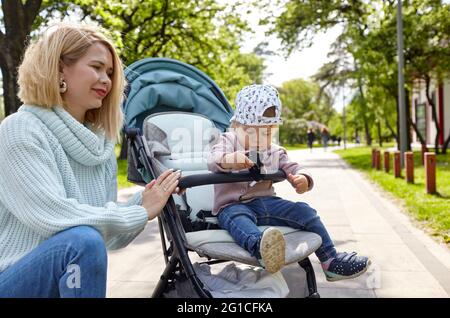 Mamma e figlia sulla natura a piedi presso il parco primaverile. Adorabile bambina in abiti comuni seduta in carrozzina blu. Bambino in buggy Foto Stock