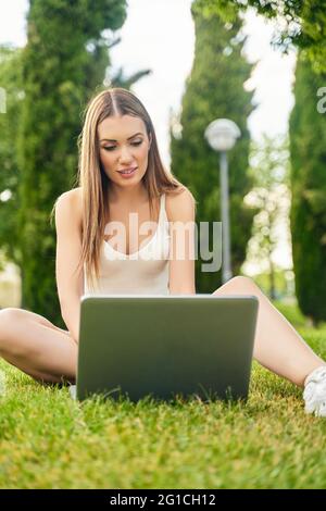 Felice giovane donna, seduta sull'erba, chiacchierando con la famiglia usando il laptop, sorridendo guardando la telecamera sullo sfondo del giardino pubblico. Concetto di persone e tecnologia. Foto di alta qualità Foto Stock