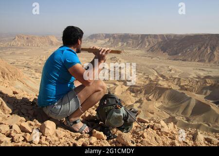 Escursionista, adulto anziano (60s) gioca flauto pastorello di legno, Monte Nishpe, Nachal Ashosh, Nachal Reim e letti asciutti del fiume sono dietro. Meyshar, deserto di Negev. Foto Stock