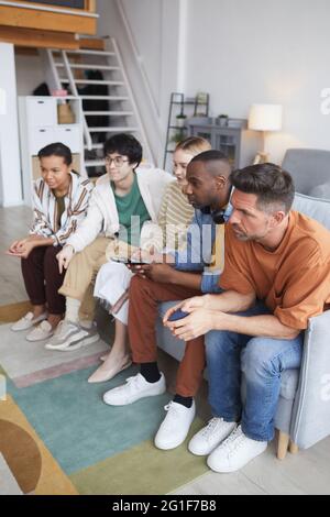 Visione verticale a tutta lunghezza di un gruppo di amici che guardano la TV a casa insieme mentre si siede in fila sul divano Foto Stock