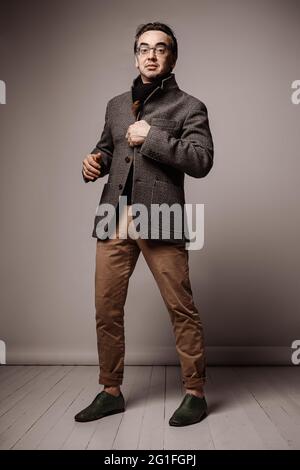 Hipster sorridente ottimista e di stile per adulti in pantaloni e sciarpa nera si alza sbottonando la sua giacca a scacchi Foto Stock