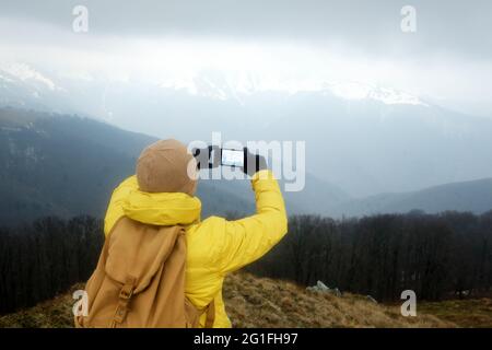 Tourist in giacca gialla con zaino facendo selfie in primavera neve montagne. Concetto di viaggio. Fotografia di paesaggio Foto Stock