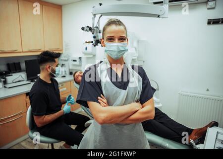 Infermiere caucasica che indossa una maschera chirurgica in piedi con le braccia incrociate in ufficio dentista Foto Stock