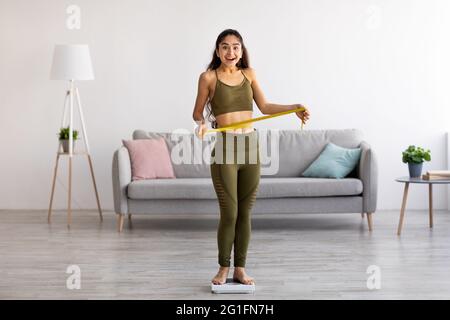 Ritratto a tutta lunghezza di una donna indiana sottile che misura la vita mentre si trova in piedi su una bilancia a casa, a tutta lunghezza Foto Stock
