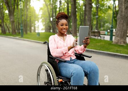 Felice giovane donna con disabilità in sedia a rotelle che utilizza un tablet per le comunicazioni online all'aperto Foto Stock