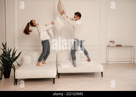 giovane coppia in maschere mediche che lotta con i cuscini mentre si divertono a casa Foto Stock