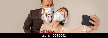 sposa in maschera medica prendendo selfie con groom isolato su grigio, banner Foto Stock