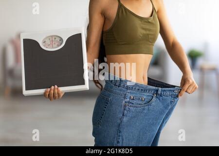 Closeup di donna indiana in jeans oversize che tengono scale, mostrando i risultati del programma di perdita di peso o liposuzione a casa Foto Stock