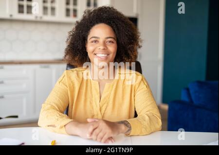 Ritratto di allegra donna afroamericana seduta in interni all'ufficio di casa. Femmina sorridente con pelle scura e acconciatura riccia indossata in blusa gialla guardando la fotocamera amichevole Foto Stock