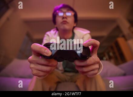 Vista ad angolo basso del giovane asiatico che tiene il gamepad e che gioca ai videogiochi a casa illuminata da luce viola, spazio di copia Foto Stock