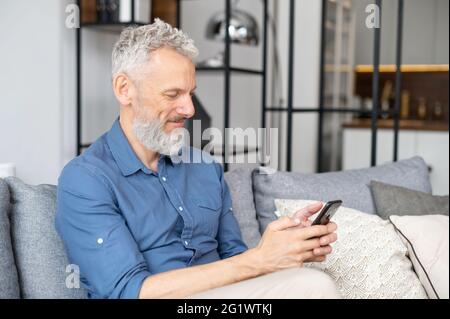 Bell'uomo maturo che invia messaggi sullo smartphone, seduto sul comodo divano con un telefono cellulare nelle mani, SMS, scorrimento e navigazione sul Web Foto Stock