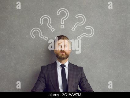 Uomo d'affari che pensa alla risposta guardando in su a parecchi punti di domanda sopra la sua testa Foto Stock