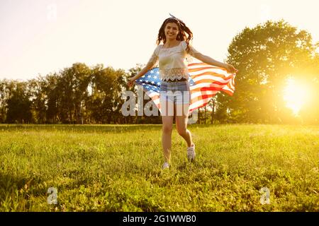 Giornata dell'indipendenza USA. Ragazza con bandiera americana corre sull'erba in un parco estivo al tramonto. Foto Stock