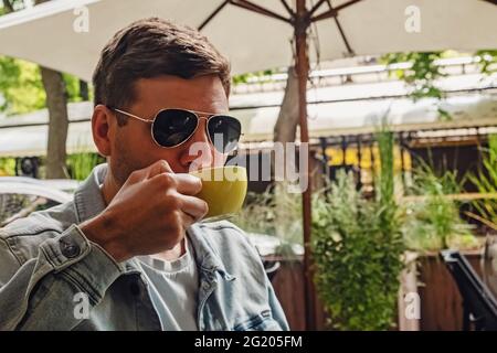 Uomo in occhiali da sole e giacca in denim seduto all'esterno nel caffè bere caffè Foto Stock