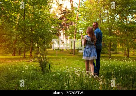 Coppia a piedi nella foresta primaverile. Giovane uomo e donna che soffia dandelioni al tramonto. Persone che godono della natura Foto Stock
