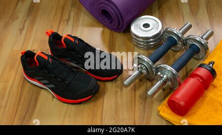 Set da palestra con paio di scarpe sportive nere, pesi per esercizi, asciugamano giallo e bottiglia rossa con acqua sul pavimento in legno Foto Stock