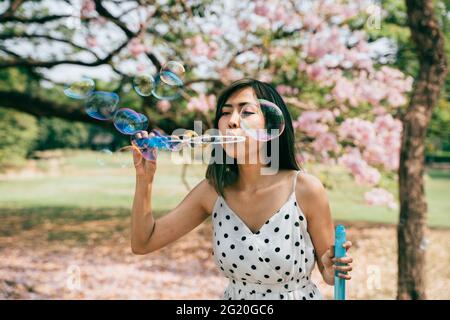 Sud-est asiatico etnico 20s donna soffia bolle d'aria nel parco giovane ragazza felice indossa un abito elegante con gli alberi sullo sfondo. Attività estive all'aperto nel fine settimana - con spazio per fotocopie Foto Stock