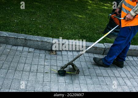 Un operaio in tute con un trimero in mano taglia l'erba sul prato e sentieri nel parco. Cura del territorio. Foto Stock