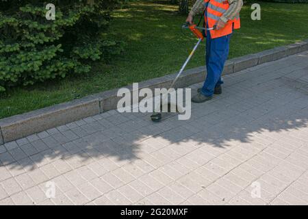 Un operaio in tute con un trimero in mano taglia l'erba sul prato e sentieri nel parco. Cura del territorio. Foto Stock