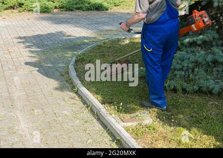 Un operaio in tute con un trimero in mano taglia l'erba sul prato e sentieri nel parco. Cura del territorio. Foto Stock