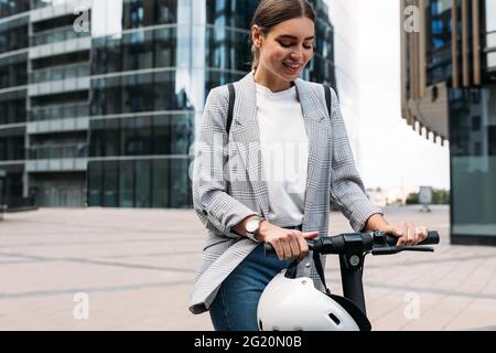 Donna bionda sorridente che regge un manubrio di un e-scooter. Donna in abbigliamento formale in piedi su scooter elettrico push pronto a guidare. Foto Stock